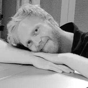 Black and white photo of a man with blond hair resting his head on his crossed arms on a table, smiling gently.